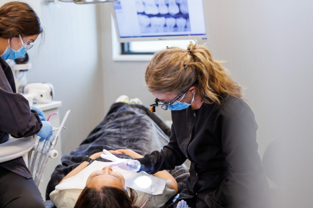 Dentist and dental assistant working on a woman's teeth while she lays in a chair with a blanket on her.