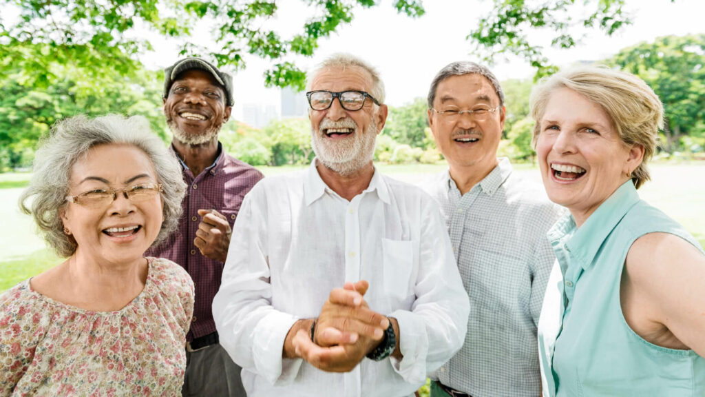 Group of diverse senior adults smiling and laughing outside underneath a tree on a sunny day.