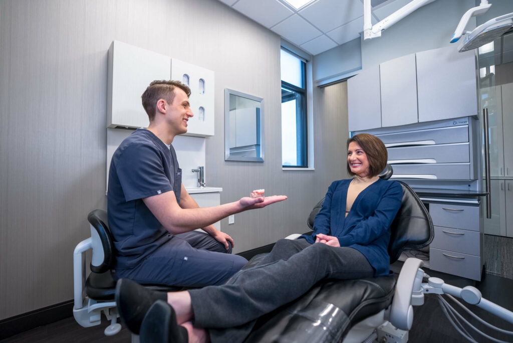 Male dentist holding a model dental implant in hand for woman in dental chair.