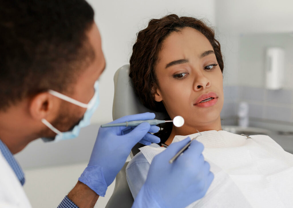 Scared black woman looking with fear at dental tools in doctor hands, denal anxiety