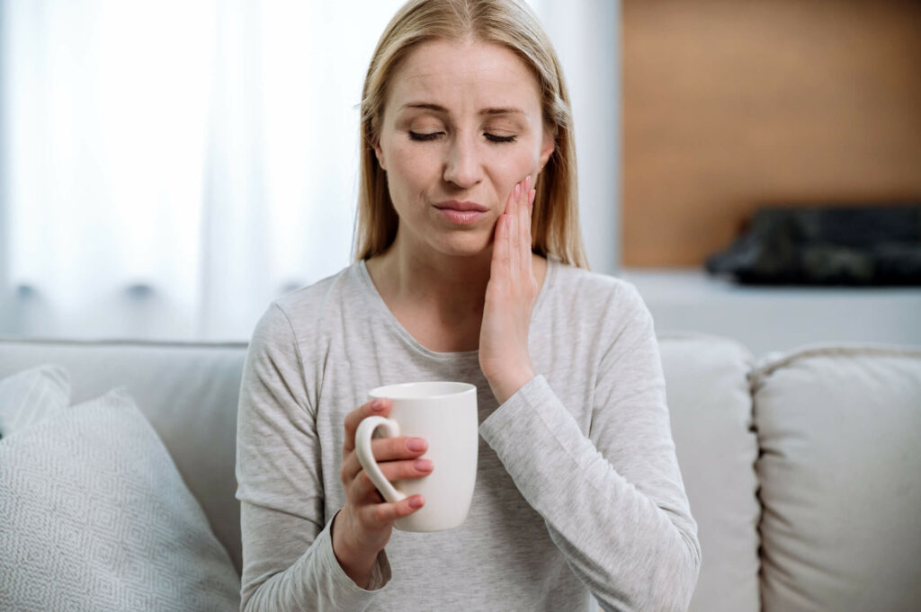 Portrait of sad woman sitting on sofa, holding cup, feeling pain while drinking hot tea or cold beverage. Sensitive teeth.