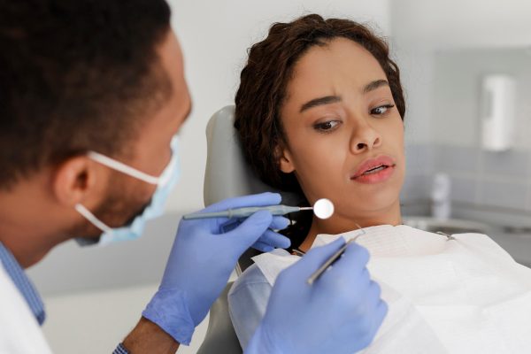 Scared black woman looking with fear at dental tools in doctor hands, denal anxiety