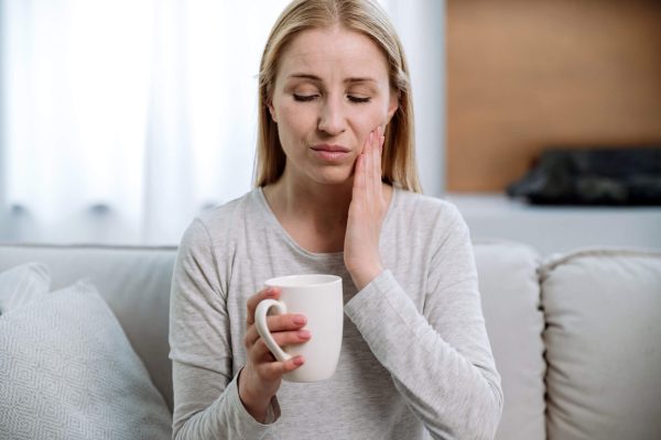 Portrait of sad woman sitting on sofa, holding cup, feeling pain while drinking hot tea or cold beverage. Sensitive teeth.